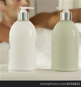 Close-up of two bottles of moisturizer with a young man in a bathtub behind it