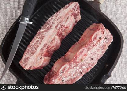 Close-up of two pieces of fresh marbled beef with sea salt and black pepper, knife on a cast-iron grill pan.