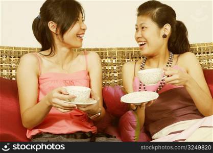 Close-up of two young women holding tea cups on a couch and smiling