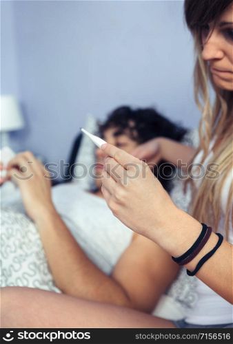 Close up of unrecognizable woman looking temperature in thermometer with sick man lying on bed in the background. Sickness and healthcare concept.. Woman looking temperature of sick man in thermometer