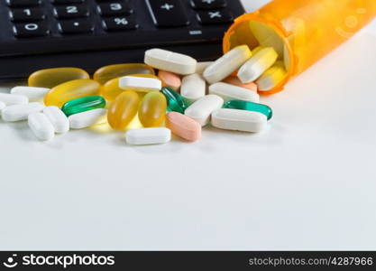 Close up of various medicine pills and tablets spilling out of plastic bottle in front of partial calculator on white background