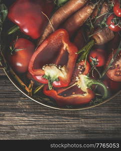 Close up of various raw red and orange summer vegetables in bowl at rustic wooden kitchen table. Halved bell pepper, carrots and tomatoes. Fresh and healthy garden harvest. Top view.