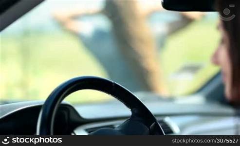 Close up of woman&acute;s hand slowly sliding on car&acute;s steering wheel. Blurred long haired blonde standing on the green field seen through the windscreen