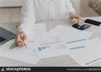 Close up of woman calculating monthly expenses, managing company budget, sitting at a desk full of documents with statistics. Female counting finances, taxes before online paying at workspace.. Female analyzing company statistics, calculates expenses, taxes before paying at workspace, close-up