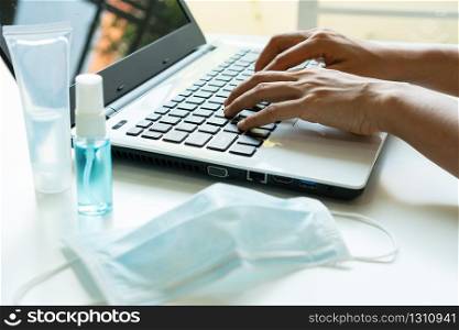 Close up of woman hands using laptop with sanitizer, alcohol spray, surgical face mask on desk at office. Protection against infectious virus, bacteria and germs concept