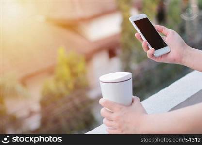 Close up of woman hands using smartphone and holding thermos mug, copy space with sunlight