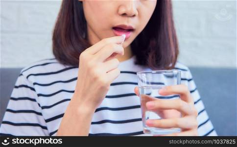 Close up of woman taking white pill in mouth and drinking water in glass on sofa in house, feels like sick. Health care concept.