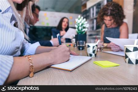 Close up of woman writing notes in a business meeting with teamwork sitting around of table in company headquarters. Woman writing notes in a meeting with teamwork