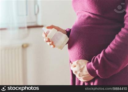Close up of young mother holding a baby bottle on her pregnant tummy, Caucasian, magenta dress