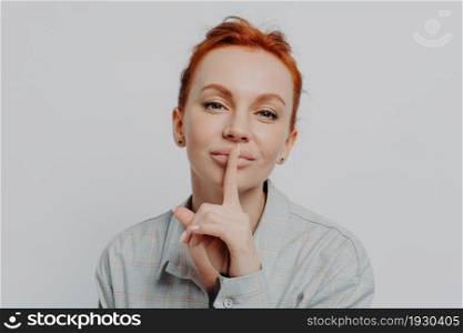 Close up of young redhead woman holding finger on lips, asking to be quiet, keep something in secret, isolated on grey studio background, caucasian female showing shh shush gesture. Caucasian female showing shh shush gesture, isolated on grey studio background