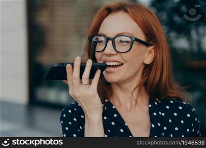 Close up of young smiling red-haired business woman holds mobile phone and using virtual digital voice assistant while standing outdoors, happy female talks on speakerphone on smartphone. Smiling red-haired business woman using virtual digital voice assistant while standing outdoors