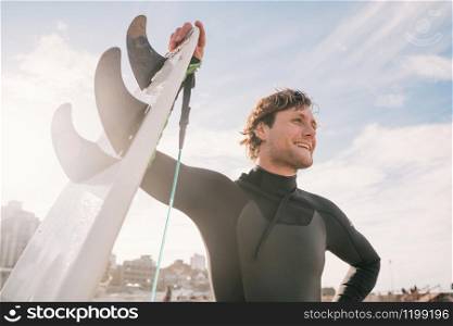 Close-up of young surfer standing at the beach with his surfboard and wearing black surfing suit. Sport and water sport concept.