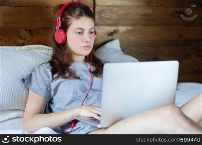 Close-up of young woman using laptop and headphones in a comfortable bed at home.