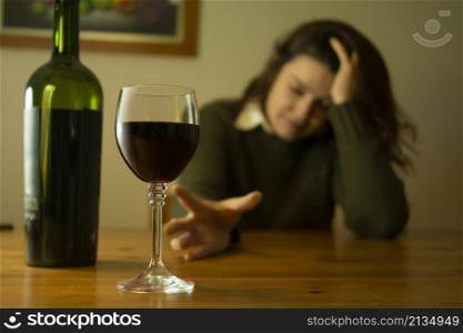 Close-up on depressed woman with green jacket sitting alone at a table trying to reach glass of red wine with her hand. Close-up of a woman with a green jacket sitting alone at a table trying to reach a glass of red wine with her hand