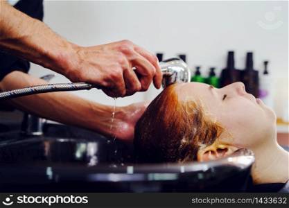 Close up on hand of adult caucasian unknown man male hairdresser holding shower head at the hairdressing salon while washing hair of unknown woman female black tub side view