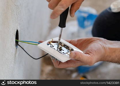 Close up on hands of caucasian man electrician holding screwdriver working on the plug electric on residential electric system installing white AC power socket on gray wall at home repair close up