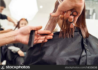 Close up on hands of unknown man male hairdresser holding scissors and comb cutting hair of unknown caucasian woman female girl customer having haircut at the salon back view