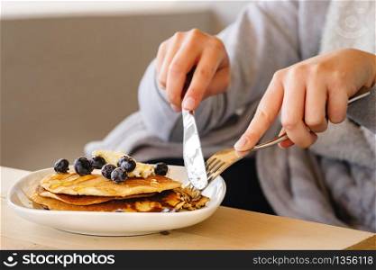 Close up on midsection female hands of unknown young woman girl holding knife and fork slicing pancakes with blueberries and maple syrup and butter while sitting by the table