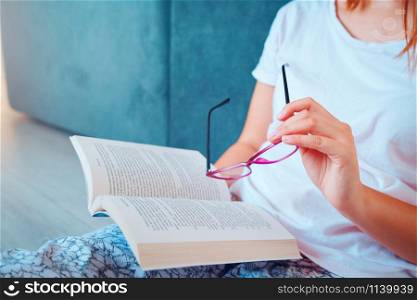 Close up on midsection of girl or woman holding a book and glasses while reading or study sitting on the floor at home