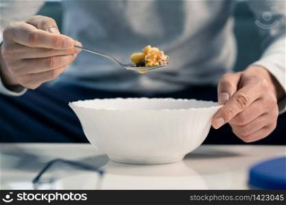 Close up on midsection of unknown caucasian man hand holding the spoon above white bowl on the table at home sitting eating muesli mix of rolled oats nuts seeds and dried fruit front view breakfast