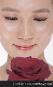 Close-up on the face of smiling beautiful woman looking down at a red rose, studio shot