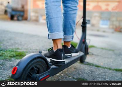 Close up on woman legs feet standing on the electric kick scooter on the pavement wearing jeans and sneakers in summer day back view