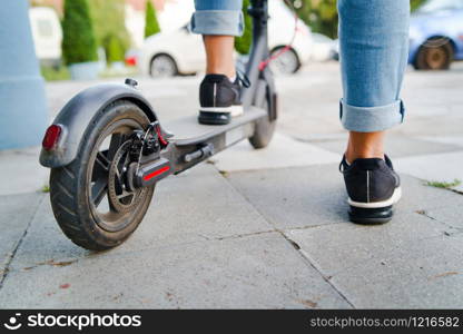 Close up on woman legs feet standing on the electric kick scooter on the pavement wearing jeans and sneakers in summer day back view