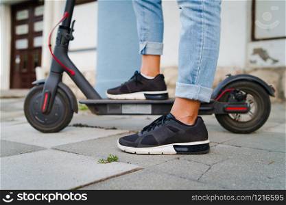 Close up on woman legs feet standing on the electric kick scooter on the pavement wearing jeans and sneakers in summer day side view riding