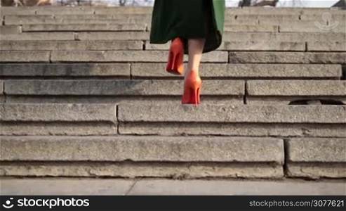 Close up perfect woman&acute;s legs in orange high heel shoes walking upstairs on stone staircase in the city. Low section. Back view of female legs in high heels wearing emerald green coat climbing up the stairs of business center. Slow motion.