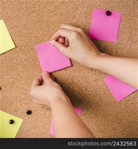 close up person fixing pink adhesive note with thumbtack corkboard