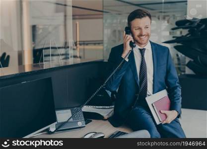 Close-up photo of attractive well-groomed confident businessman in formal wear, speaking on his office desk phone while sitting on top of table, holding laptop and agenda, happy to hear good news. Businessman in formal wear, speaking on his office desk phone while sitting on top of table