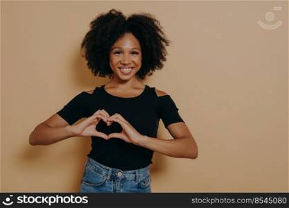 Close up photo of beautiful amazing dark skinned lady showing with hands sweetheart symbol, love message gesture, wears casual black t-shirt isolated over dark beige studio background with copy space. Close up photo of beautiful amazing dark skinned lady showing with hands sweetheart symbol