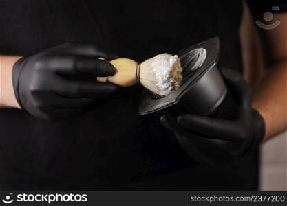 Close up photo of hairstylist hands holding bowl of shaving foam and wooden shaving brush. man He in black glovers is standing indoor barbershop . Shaving accessories. Close up photo of hairstylist hands holding bowl of shaving foam and wooden shaving brush. man He in black glovers is standing indoor barbershop . Shaving accessories.