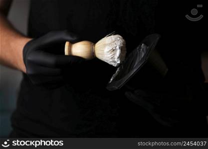 Close up photo of hairstylist hands holding bowl of shaving foam and wooden shaving brush. man He in black glovers is standing indoor barbershop . Shaving accessories. Close up photo of hairstylist hands holding bowl of shaving foam and wooden shaving brush. man He in black glovers is standing indoor barbershop . Shaving accessories.