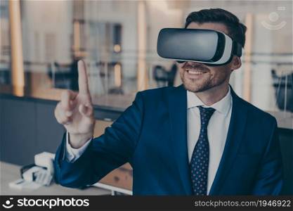 Close-up photo of smiling young bearded finacier in formal suit enjoying spending time in virtual reality with VR headset glasses, pointing with forefinger up in air. Cyberspace experience concept. Smiling young finacier in formal suit enjoying spending time in virtual reality with VR headset