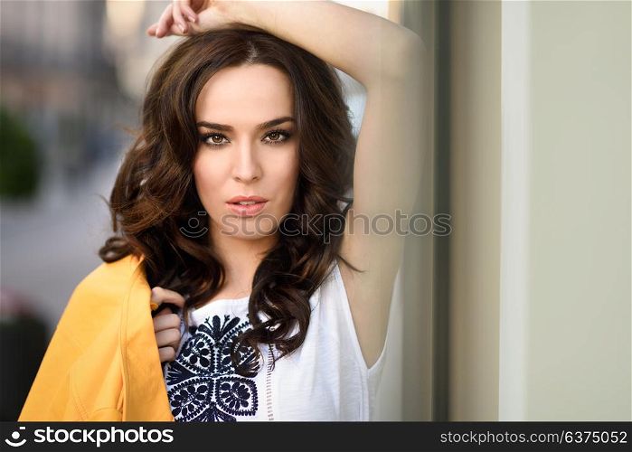 Close-up portraig of young brunette woman, model of fashion, wearing orange modern jacket in urban background.