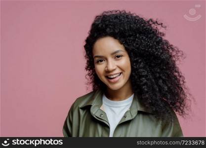 Close up portrair of smiling young female model has curly hair, healthy skin, looks directly at camera, expresses positive emotions, enjoys being photographed, dressed in fashionable clothes