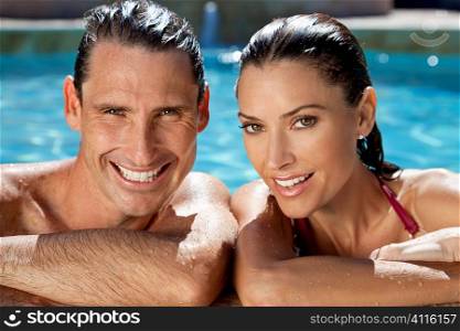 Close up portrait of a beautiful happy man and woman couple resting on their hands at the side of a sun bathed swimming pool smiling with perfect teeth.