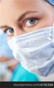 Close up portrait of a blue eyed female doctor wearing scrubs and a surgical mask, her colleague is out of focus in the distant background.