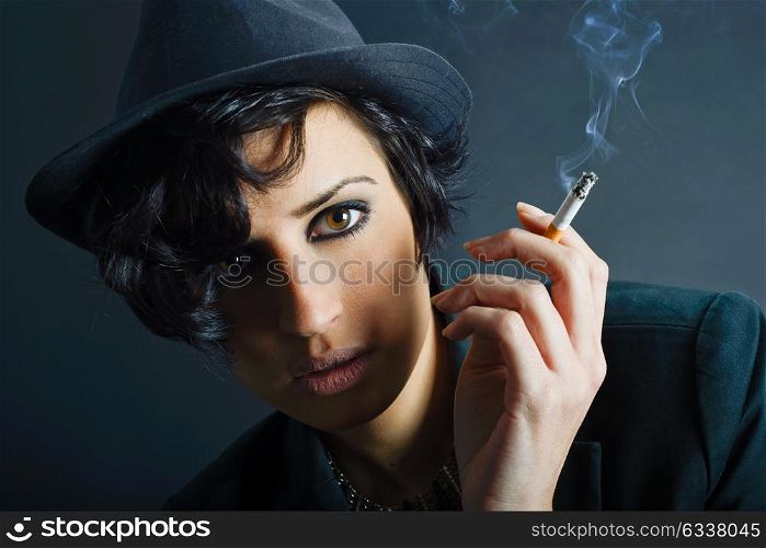 Close-up portrait of a brunette woman smoking a cigarette on black background wearing a hat