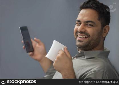 Close-up portrait of a cheerful young man looking at camera with holding coffee mug and mobile in hands