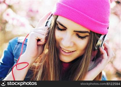 Close up portrait of a funny young swag girl on lush garden listening to music in earphones from smart phone mp3 player. Outdoors, lifestyle.