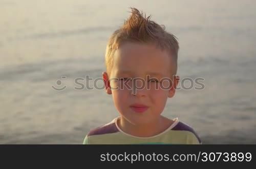Close-up portrait of a happy little blond boy with mohawk on the background of wavy sea, shot in evening sunlight