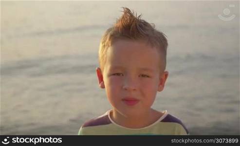Close-up portrait of a happy little blond boy with mohawk on the background of wavy sea, shot in evening sunlight