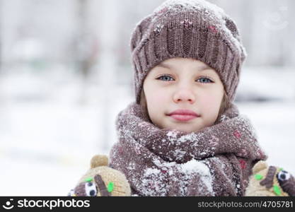 Close-up portrait of a little girl in brown jacket and knit scarf and hat on a background of a snow park