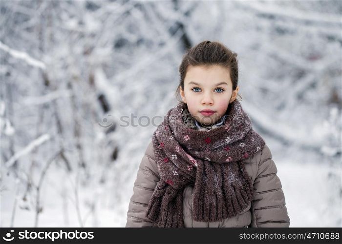 Close-up portrait of a little girl in brown jacket and knit scarf and hat on a background of a snow park