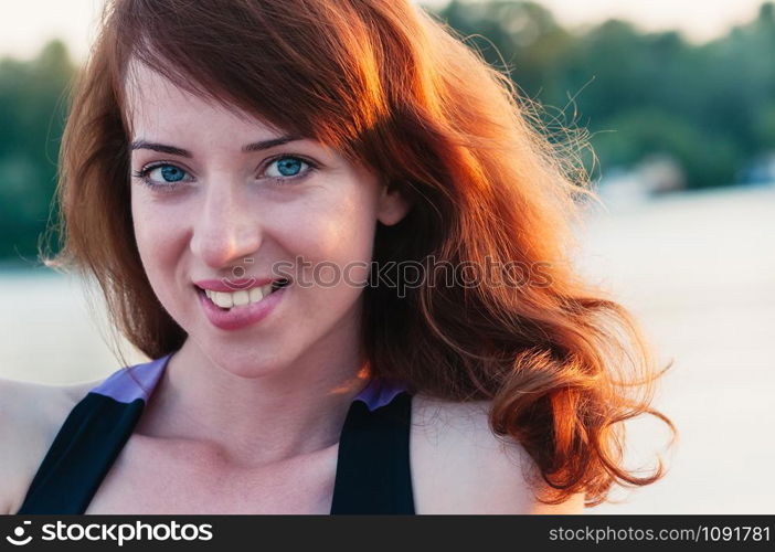 Close up portrait of a young beautiful ginger haired woman, biting her lip in a smile, white teeth, looking at camera, on water summer nature background