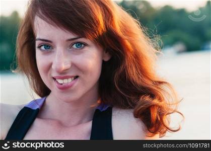 Close up portrait of a young beautiful ginger haired woman, biting her lip in a smile, white teeth, looking at camera, on water summer nature background