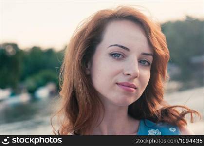 Close up portrait of a young beautiful ginger haired woman, looking at camera, blue eyes and clothes, sunset light, summer nature background, motion blur, messy hair by the wind. Street fashion