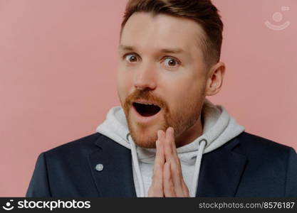 Close up portrait of amazed bearded young European man keeps mouth opened palms pressed together cannot believe in something wears sweatshirt with hood black jacket isolated over pink background